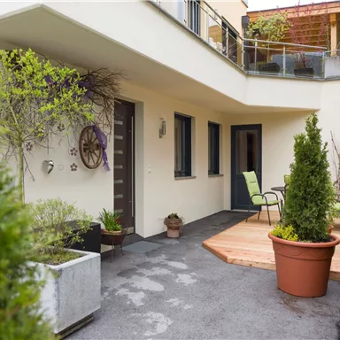 Ein einladender Eingangsbereich mit Pflanzen und einer Holzterrasse. Auf der Terrasse steht ein Stuhl, um die frische Luft zu genießen.