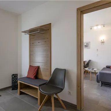 Ein moderner Eingangsbereich mit einer Holzbank und einem Stuhl. Die Wände sind hell, und es gibt einen Blick auf ein angrenzendes Zimmer.