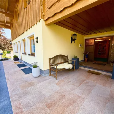 An inviting entrance area of a house with a wooden porch. Flowers and plants adorn the ground and the architecture shows alpine influences.
