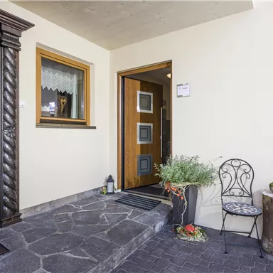 Ein einladender Eingangsbereich mit einer schönen Tür und einem gemütlichen Stuhl. Pflanzen und eine kleine Laterne verleihen dem Platz eine angenehme Atmosphäre.