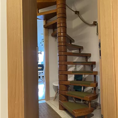 Eine helle Wendeltreppe aus Holz führt in den oberen Bereich eines Raumes. Durch die offene Tür sieht man einen einladenden Wohnbereich.