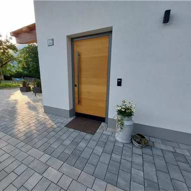 A modern wooden entrance door with a matte doormat. Next to the door, there is a flower pot with white flowers.