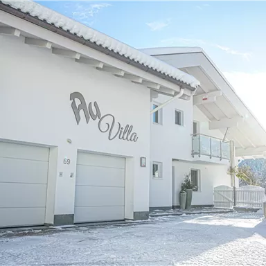Ein modernes, schneebedecktes Haus mit der Aufschrift "Aqua Villa". Die Fassade ist hell und einladend unter einem klaren Himmel.