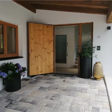 Ein einladender Eingangsbereich mit einer offenen Tür und modernen Fliesen. Auf der Terrasse stehen Pflanzen in Töpfen und eine dekorative goldene Vase.