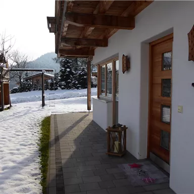 Ein gemütliches Haus im Winter mit Schnee auf dem Boden. Im Hintergrund sind Spielgeräte und Bäume zu sehen.