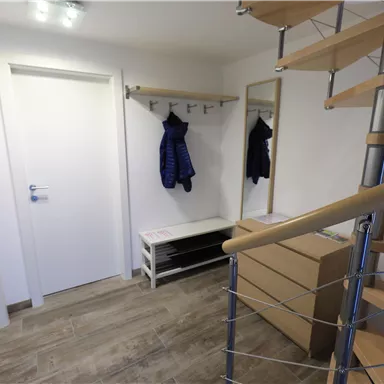 A bright hallway with a staircase, a mirror, and a coat rack. There is a bench and a closed cupboard on the wall.