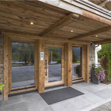 Entrance of a wood-clad building with large glass windows. The foyer is surrounded by green plants and blooming flowers.