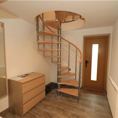 A bright entrance area with an elegant spiral staircase made of wood and metal. On the right, there is a door with glass inserts and a modern chest of drawers.