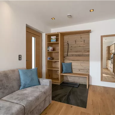 A bright entrance area with a cozy sofa and a wooden shelf. The room has a modern decor and a large mirror.