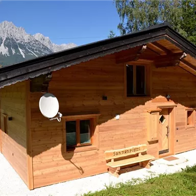 A cozy wooden house with a large garden and a beautiful mountain panorama in the background. The sun is shining on the building, which looks rustic.