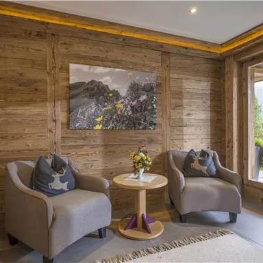 A cozy interior with wooden paneling and two beige armchairs. In the center stands a round table with a floral decoration and a large window that looks out onto a green landscape.