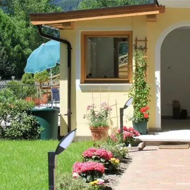 Ein einladender Eingangsbereich mit einem gepflegten Garten und bunten Blumen. Im Hintergrund sieht man einen blauen Himmel und Bäume.