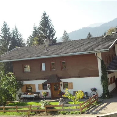 A cozy, traditional house surrounded by trees and blooming plants. Gentle hills can be seen in the background.