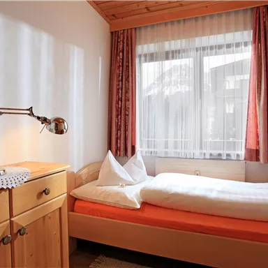 Ein kleines, gemütliches Schlafzimmer mit einem Einzelbett und einem Holzschrank. Große Fenster lassen viel Tageslicht herein und die Fenster sind mit Vorhängen geschmückt.
