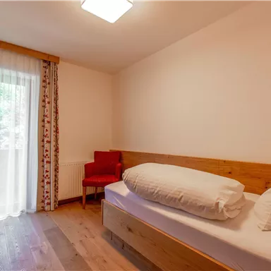 A simple room with a single bed and a red chair. Large windows provide plenty of light and a view outside.