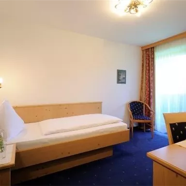 A simple and cozy single room with wooden furniture. There is a bed, a table, and a chair, as well as a window with curtains.