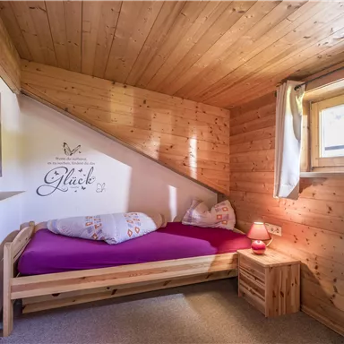 A cozy room with wooden walls and a single bed. A window lets in natural light, and there is a decorative vase on the table.