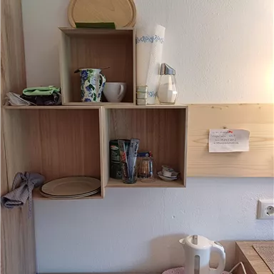 Eine Holzregal mit verschiedenen Fächern. Darin befinden sich Geschirr, eine Kaffeekanne und eine Wasserkocher auf einem Tisch.