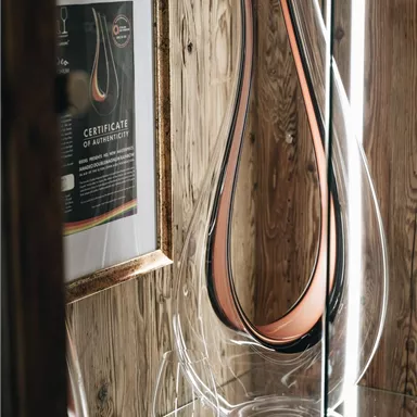 An elegant glass vase with a modern design stands in a showcase. Next to the vase, a certificate and some wine glasses can be seen.