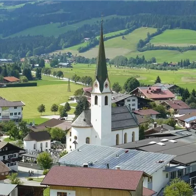 A picturesque landscape with a charming village and a characteristic church. The surroundings are green and hilly with many trees and buildings.