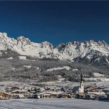 A picturesque winter landscape with snow-covered mountains and a small village. The sky is clear and blue, creating a peaceful atmosphere.