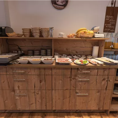A breakfast buffet with a wooden coast displaying various foods such as cold cuts, cheese, and fruit. In the background, plates and bowls are neatly arranged.