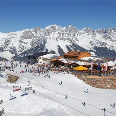 A lively ski resort with many people on the slopes. The impressive mountains surround the scene and the sky is clear and blue.