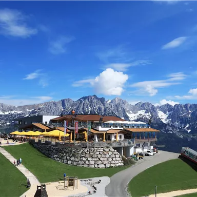 A picturesque mountain cabin with a terrace and sun umbrellas in the Alps. In the background, majestic mountains rise under a clear blue sky.