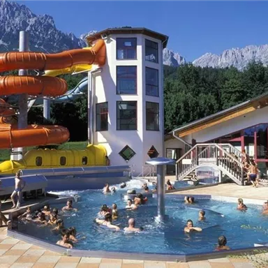 A recreational pool with a large water slide and an indoor pool. In the background, mountains can be seen, while many children and adults are having fun in the water.