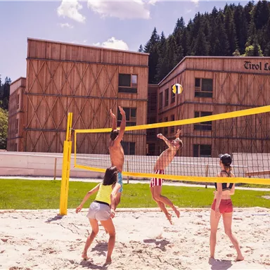 An outdoor beach volleyball game with several players. In the background, modern wooden houses and green forests can be seen.