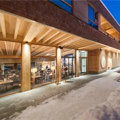 A modern building with large windows and a wooden facade, surrounded by snow. Inside, people are visible, sitting comfortably and engaging in conversation.