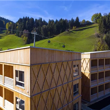 Modern wooden buildings in a hilly landscape. In the background, green meadows and trees can be seen.