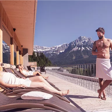 A relaxed wellness scene on a terrace in the mountains. People in bathrobes enjoy the view of the Alps.