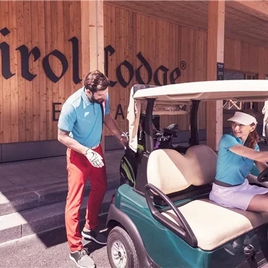 A golf course with a golf cart and two players. In the background, the Tirol Lodge sign can be seen.