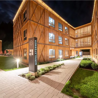 A modern wooden building at night, illuminated by warm light. The well-maintained outdoor area with green lawns invites you to linger.