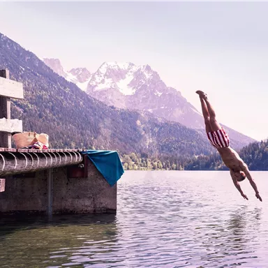 A man jumps into the water from a wooden pier. In the background, there are mountains and a clear sky.