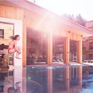 A girl jumps into a pool in front of a modern wooden building. The sun is shining, and it is a clear, beautiful day.