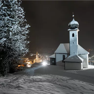 A snow-covered landscape at night with a small church and a tall church tower. In the background, the lights of a small village are glowing.
