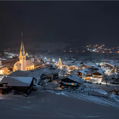 A snowy city at night with warm lights. In the background, a church with a tall tower can be seen.