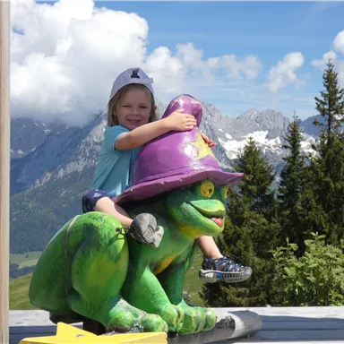 A small child sits on a colorful frog figure with a hat. In the background, there are mountains and a blue sky with clouds.