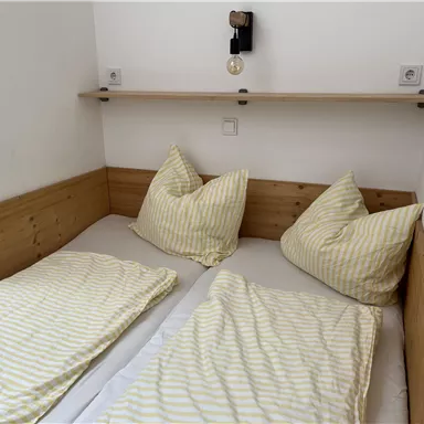 Ein einfaches Schlafzimmer mit einem Doppelbett und gestreiften Bettwaren. An der Wand hängt eine Lampe und ein Regal.
