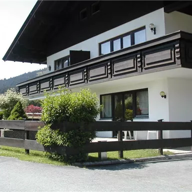 A beautiful house in alpine style with a black balcony railing. The garden is well-kept and the surroundings are surrounded by mountains.