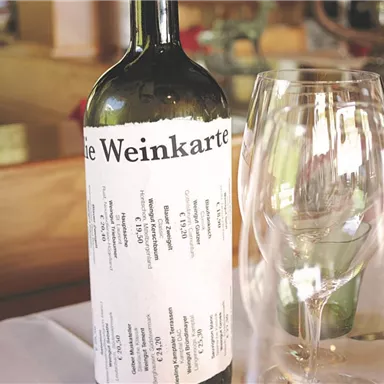 A bottle of wine is next to a glass on a table. The label on the bottle displays the wine list.