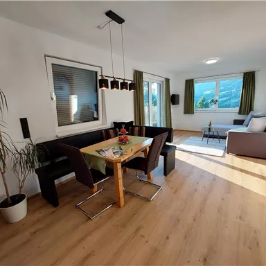 A bright and modern living room with a dining table and black seating area. Large windows let in plenty of light and offer a view outside.