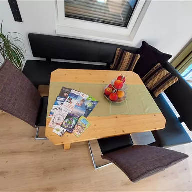 A cozy dining area with a wooden table and comfortable seating. On the table are some magazines and a decorative bowl with apples.
