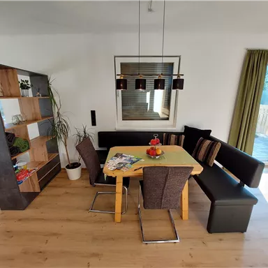 A modern dining room with a wooden table and a cozy corner sofa. The room is bright and friendly, with plants and a large window front.