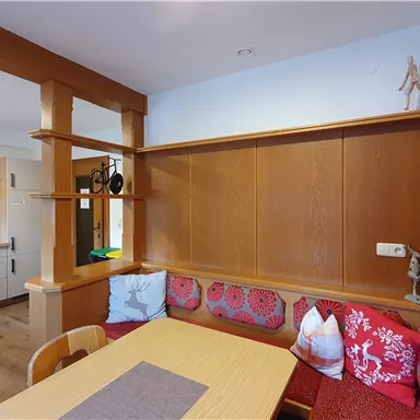 A cozy dining area with wooden paneling and colorful cushions. The open kitchen is visible in the background.