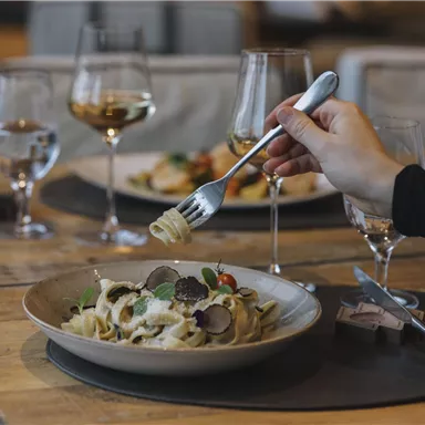 Ein Teller Pasta mit frischen Zutaten steht auf einem Holztisch. Im Hintergrund sind Weingläser und ein weiterer Teller mit Essen zu sehen.