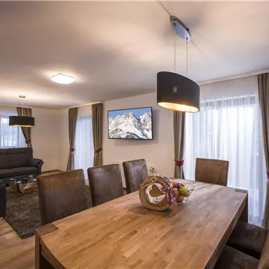 A modern living space with a large dining table and comfortable chairs. In the background, there is a sofa and a television with a mountain landscape visible.