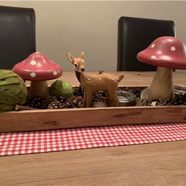 A decorative table arrangement with red mushrooms, a wooden deer, and green elements. The decoration is placed on a checkered table runner.